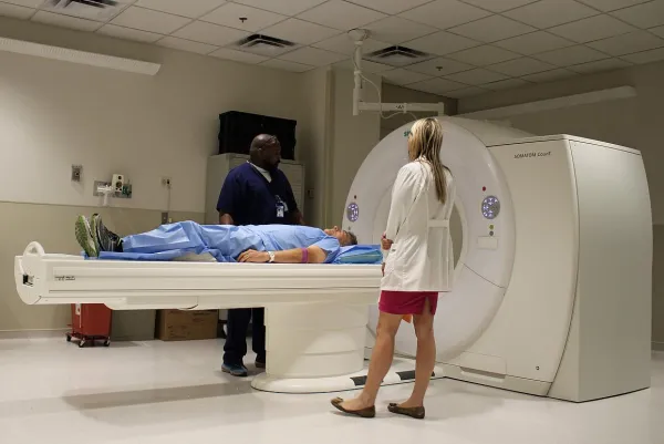 A doctor and a nurse stand on either side of a patient; the patient is laying down at the head of a CT machine.